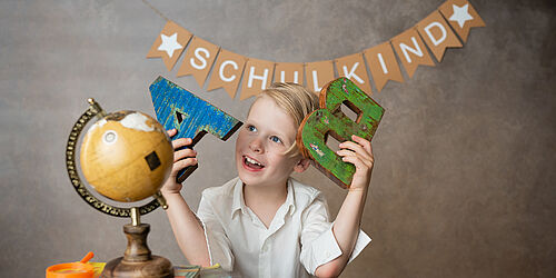 Kleiner lächelnder Junge mit Holzbuchstaben A und B in der Hand. Im Hintergrund ein Banner mit dem Wort "Schulkind".