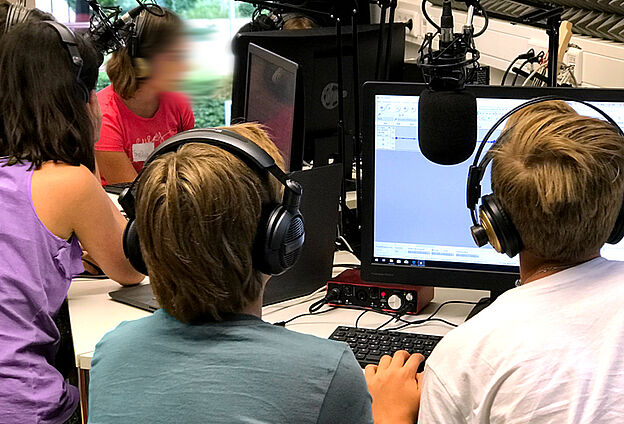 Foto Podcast-Lab in der Feierwerk Funkstation. Zu sehen sind fünf Kinder, die um Computerbildschirme und Mikros sitzen.